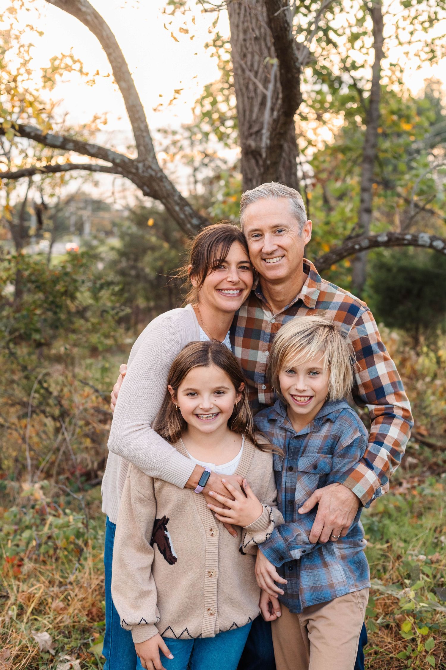 Smiling family of four hugging together in autumn woodland setting at sunset.