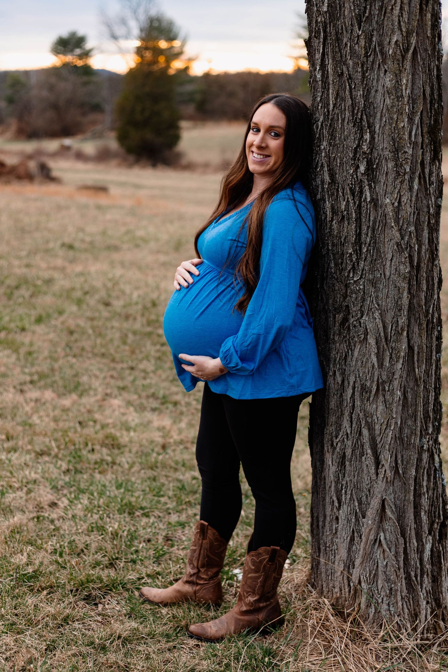 Smiling pregnant woman in blue top and boots leaning against tree during outdoor maternity session in Douglassville, PA.