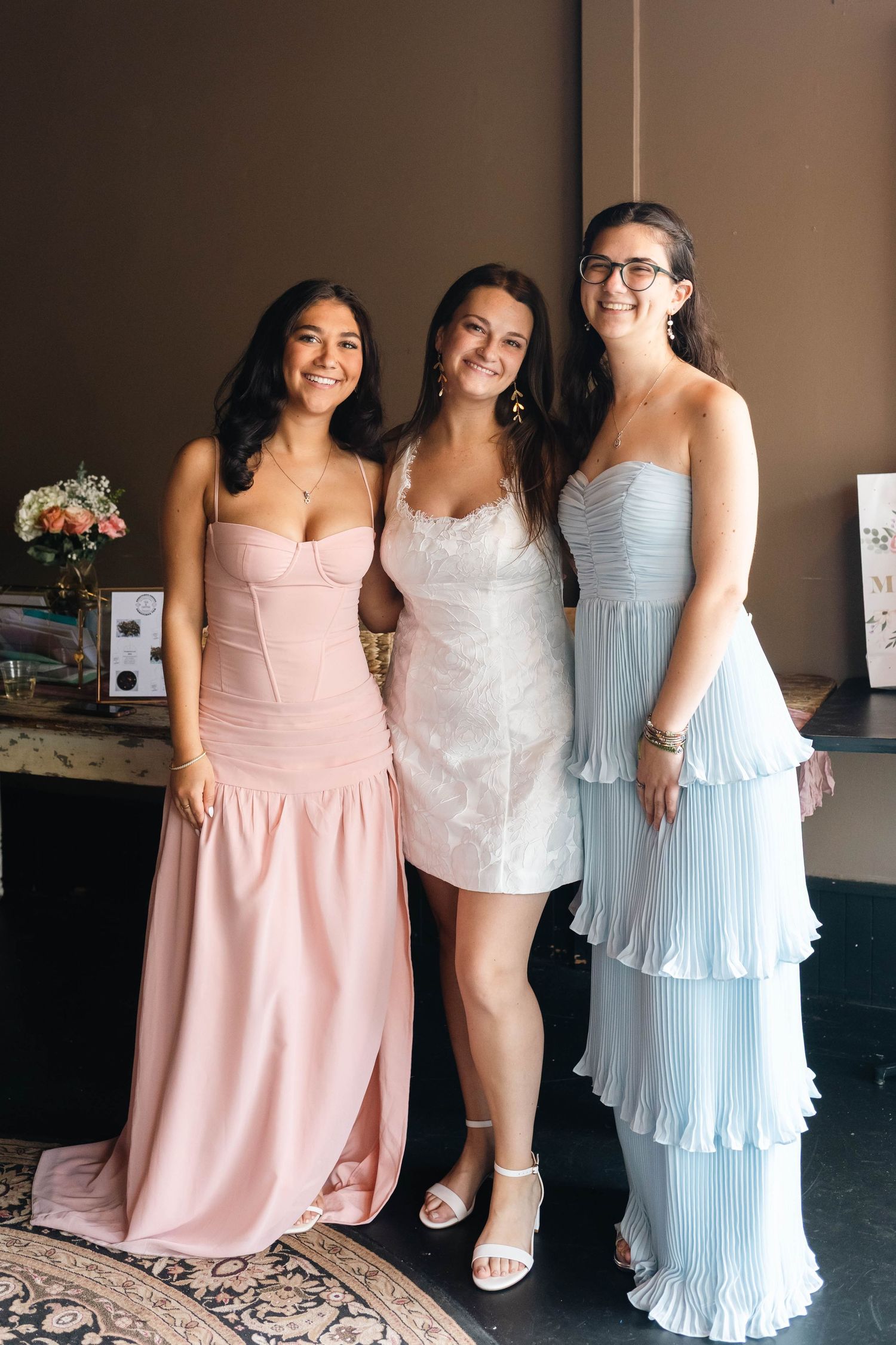 Three women posing together at wedding, wearing pink, white, and light blue formal dresses.