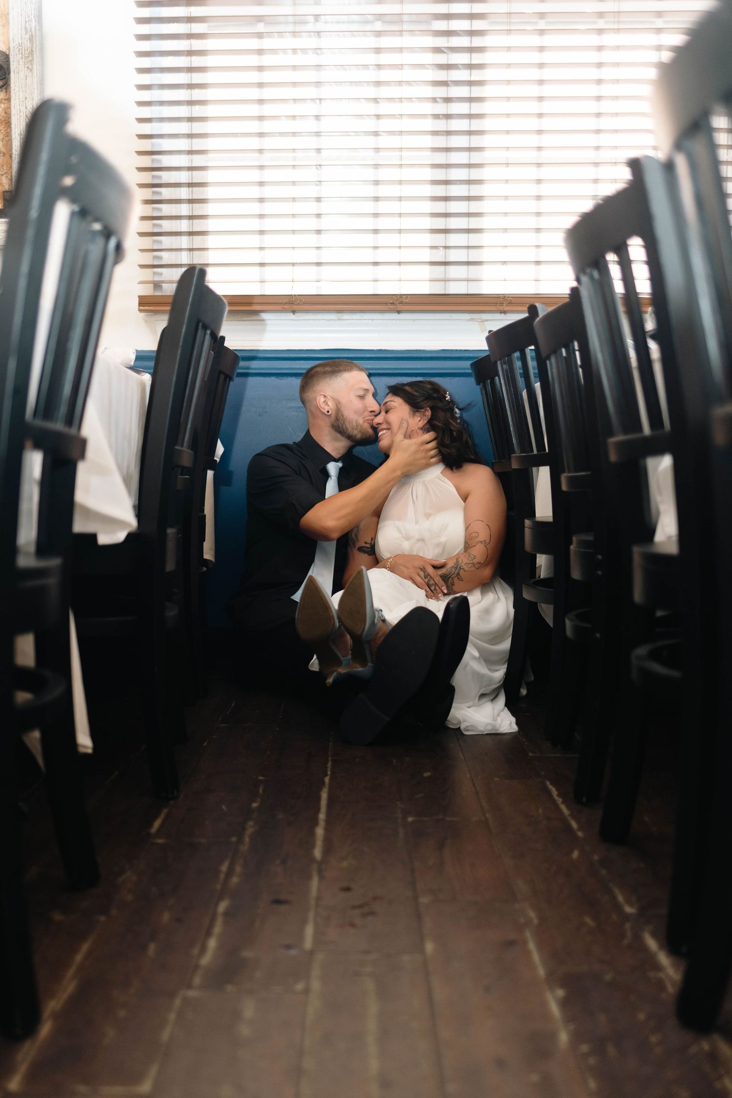 Bride and groom sharing a romantic kiss while sitting on wooden floor between rows of stacked chairs.
