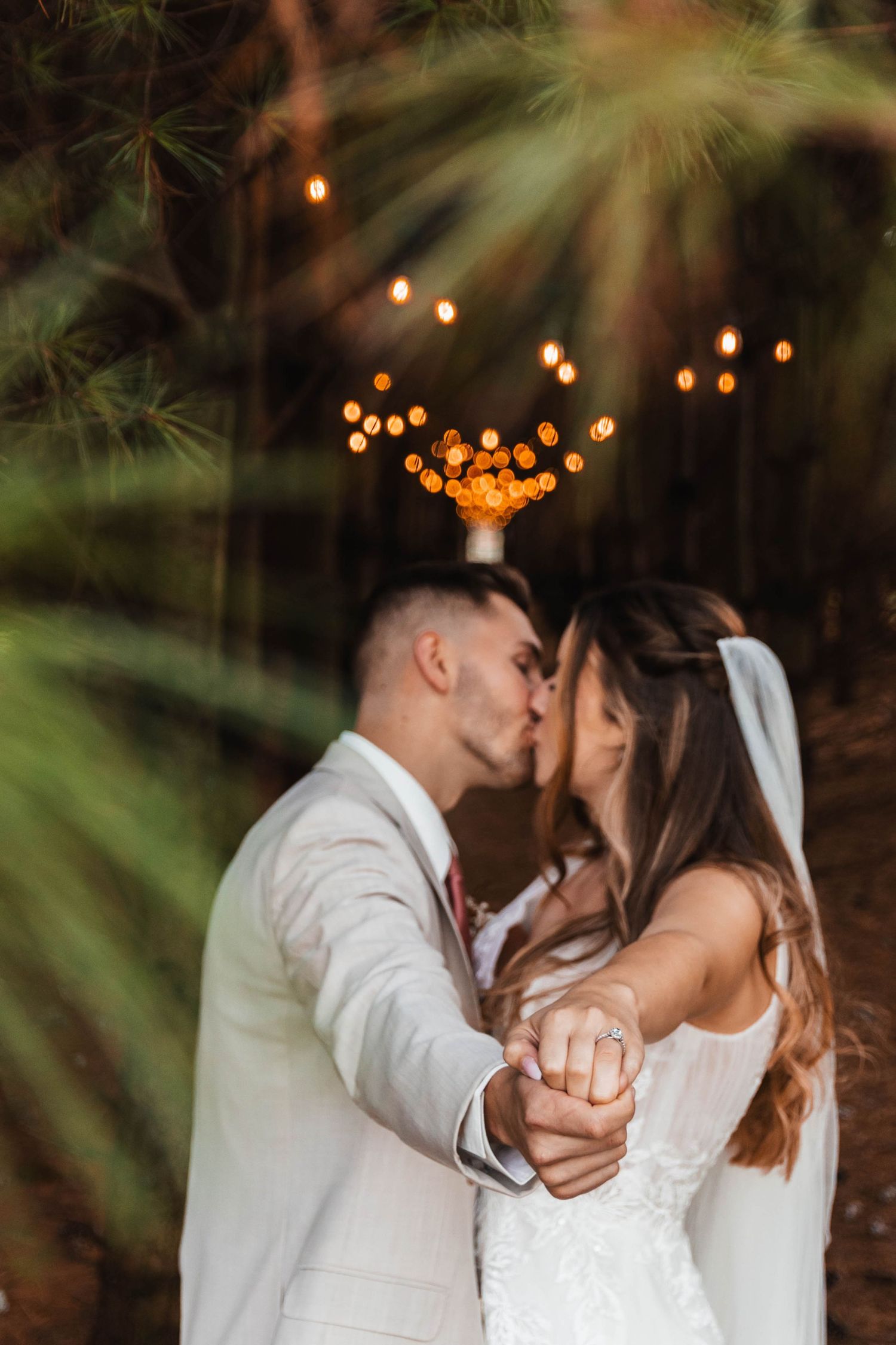 Bride and groom share a kiss while holding hands under pine trees with bokeh string lights above.