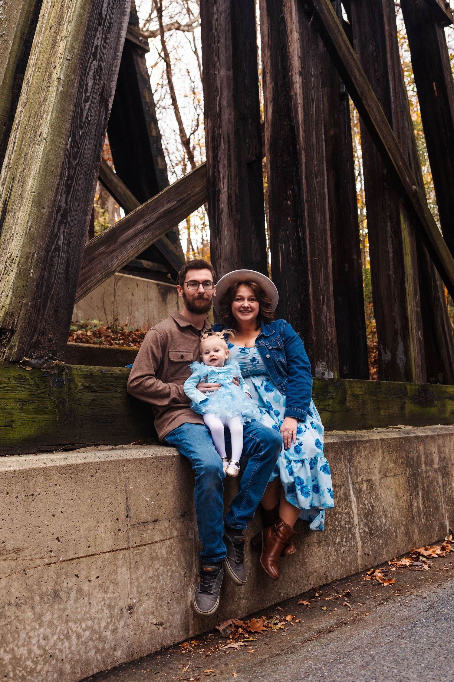 Young family with baby sits on stone wall beneath wooden trestle structure surrounded by fall foliage in Boyertown, PA.