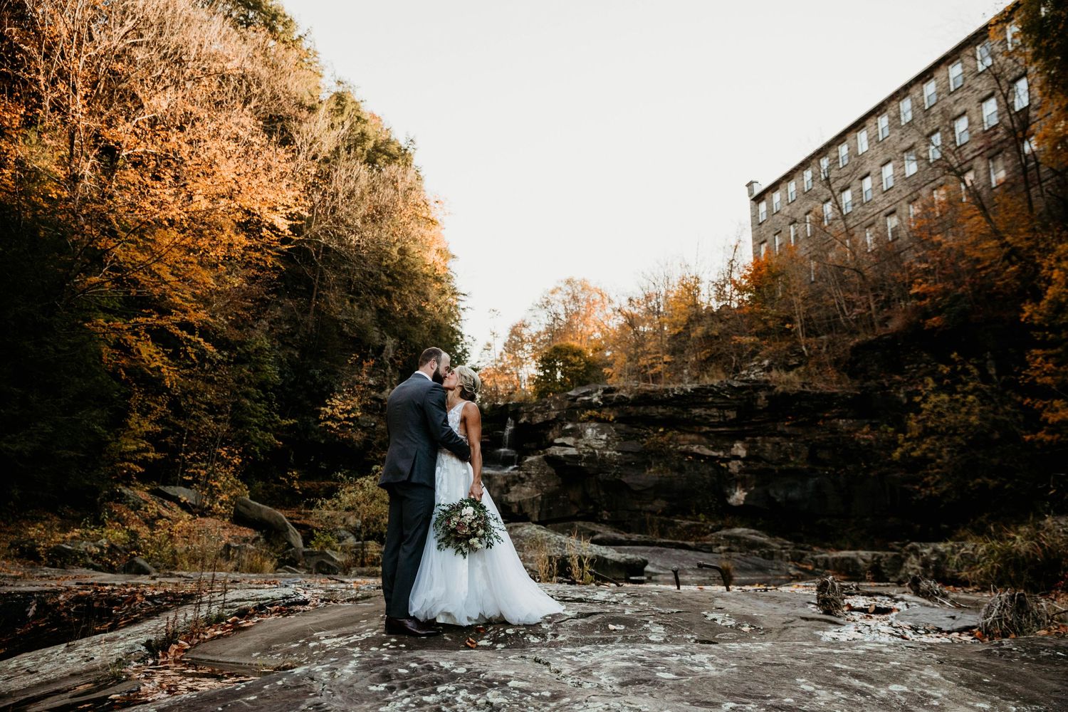 Couple sharing romantic kiss on rocky waterfall surrounded by autumn foliage near rustic mill building.