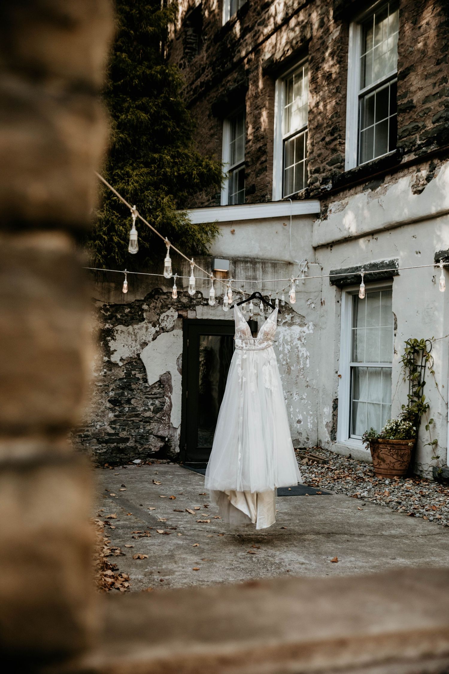 Elegant white wedding dress hanging on string lights outside a rustic stone building.