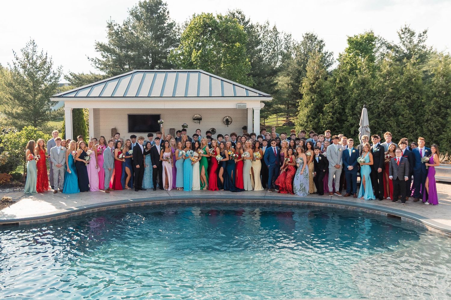 Large group of Perkiomenville prom students in formal attire posing together poolside in front of a pool house.