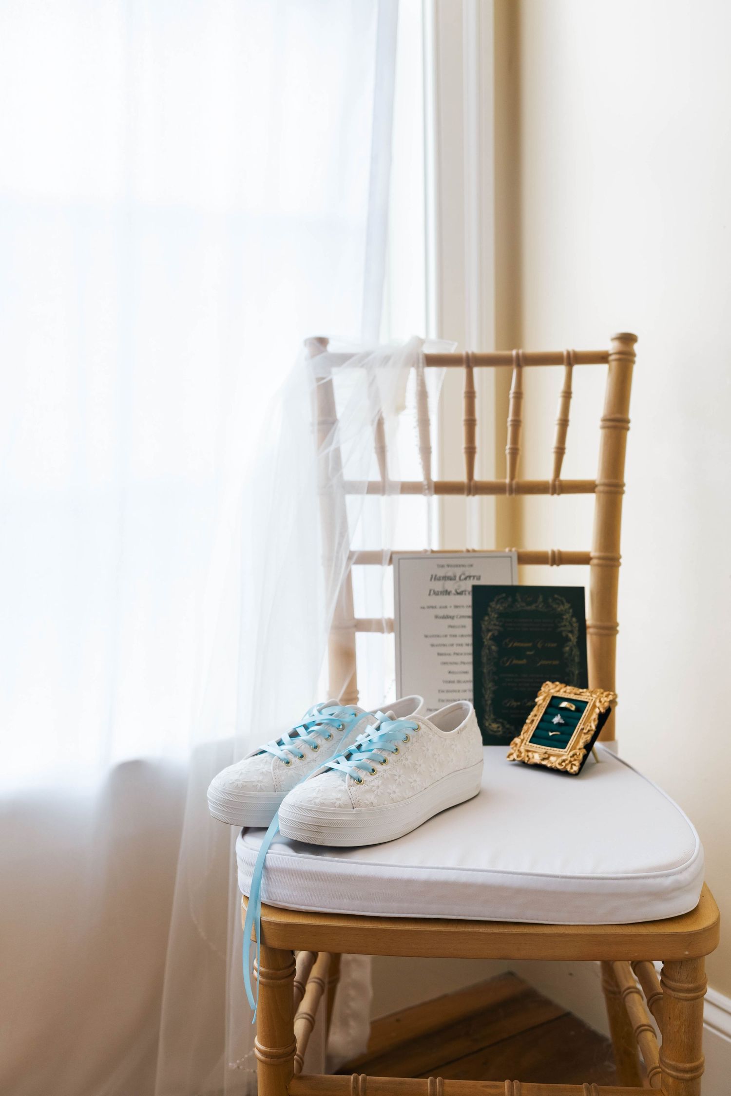 White lace sneakers with blue laces beside a wedding program and gold frame on a chiavari chair.