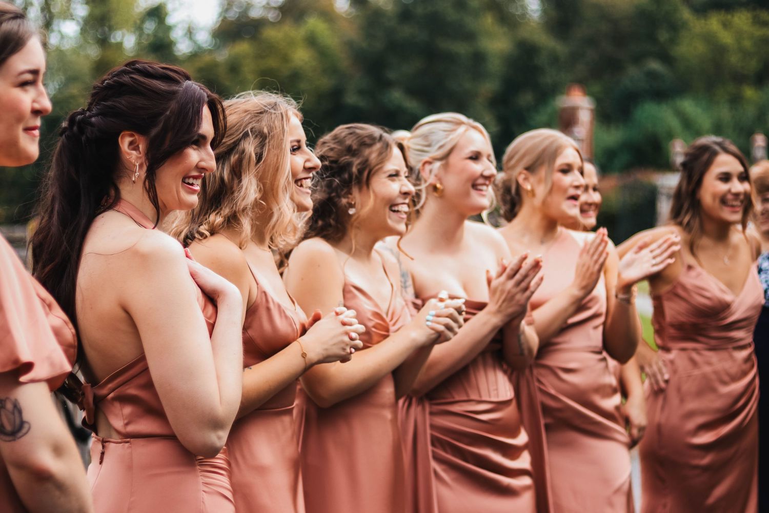 Bridesmaids in dusty rose satin dresses laugh and clap joyfully during a first look.