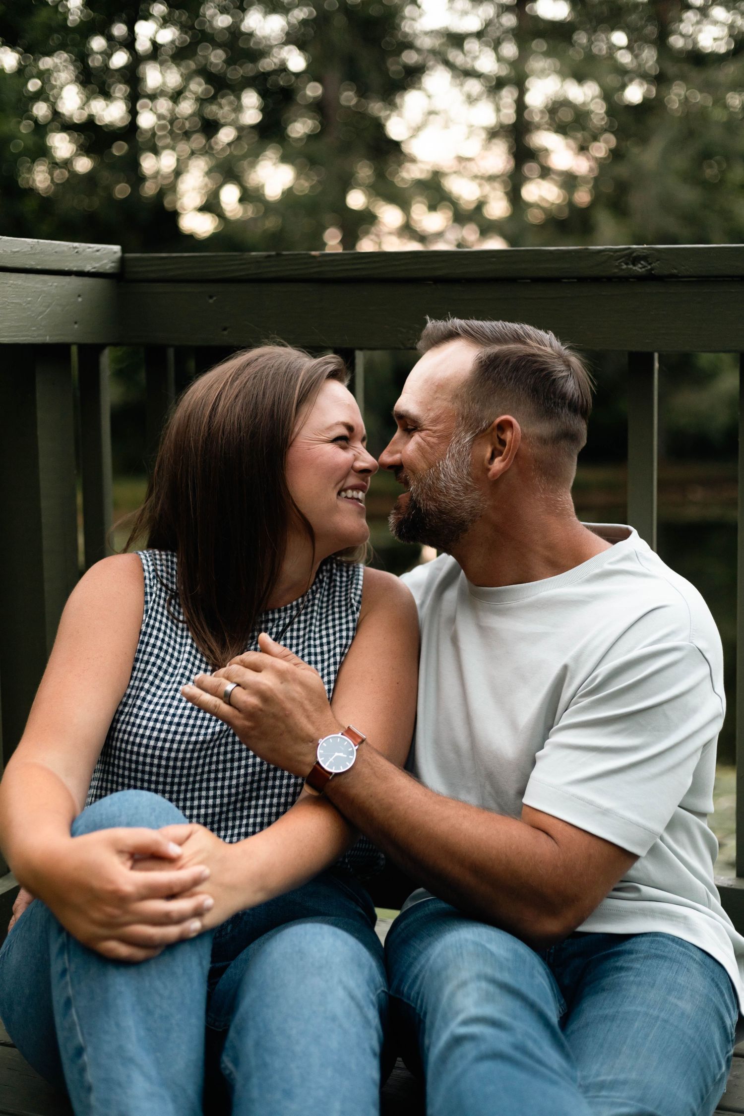 Smiling couple sitting close together on a deck, sharing a tender moment surrounded by lush green trees.