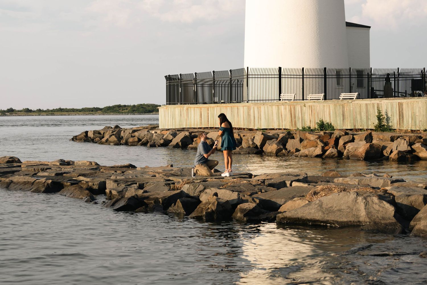 Two children explore rocky shoreline near a white lighthouse surrounded by calm waters at sunset in Barnegat, NJ.