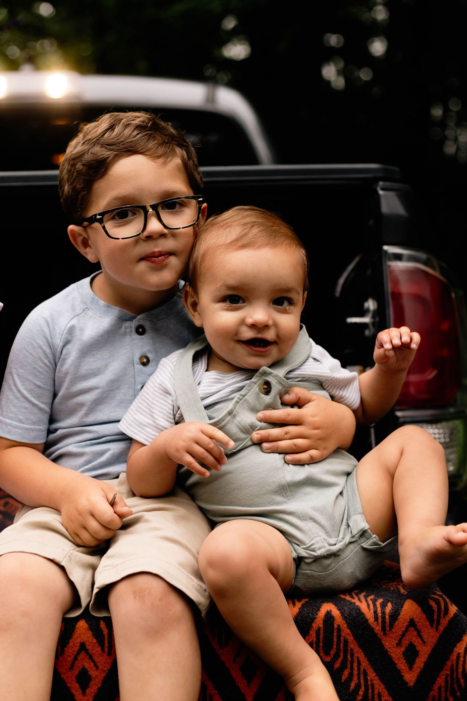 Two young brothers sitting in a truck bed, older boy with glasses hugging his baby sibling at Lockridge Park.