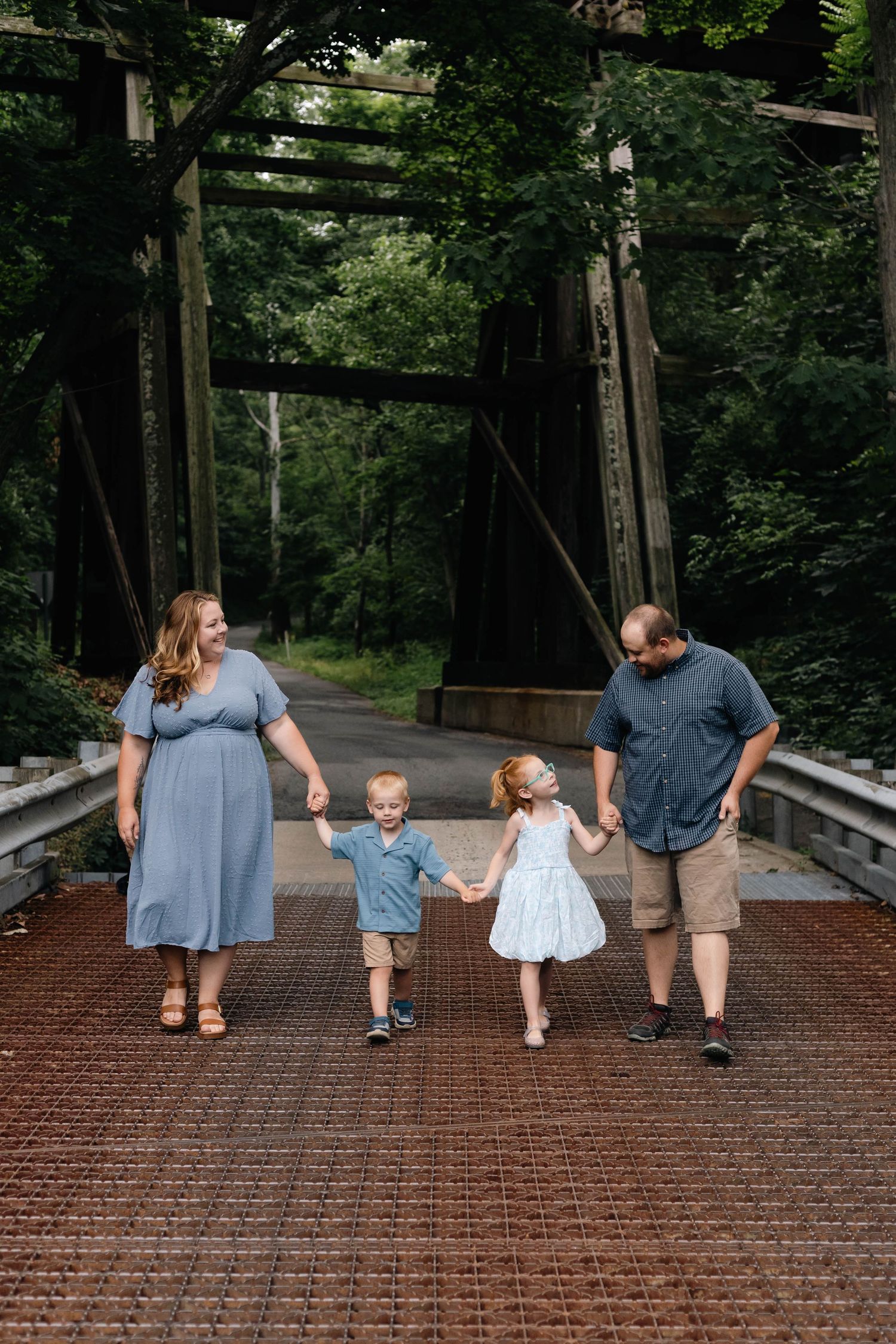 Happy family of four holds hands walking across rustic iron bridge surrounded by lush green forest in Boyertown, PA.