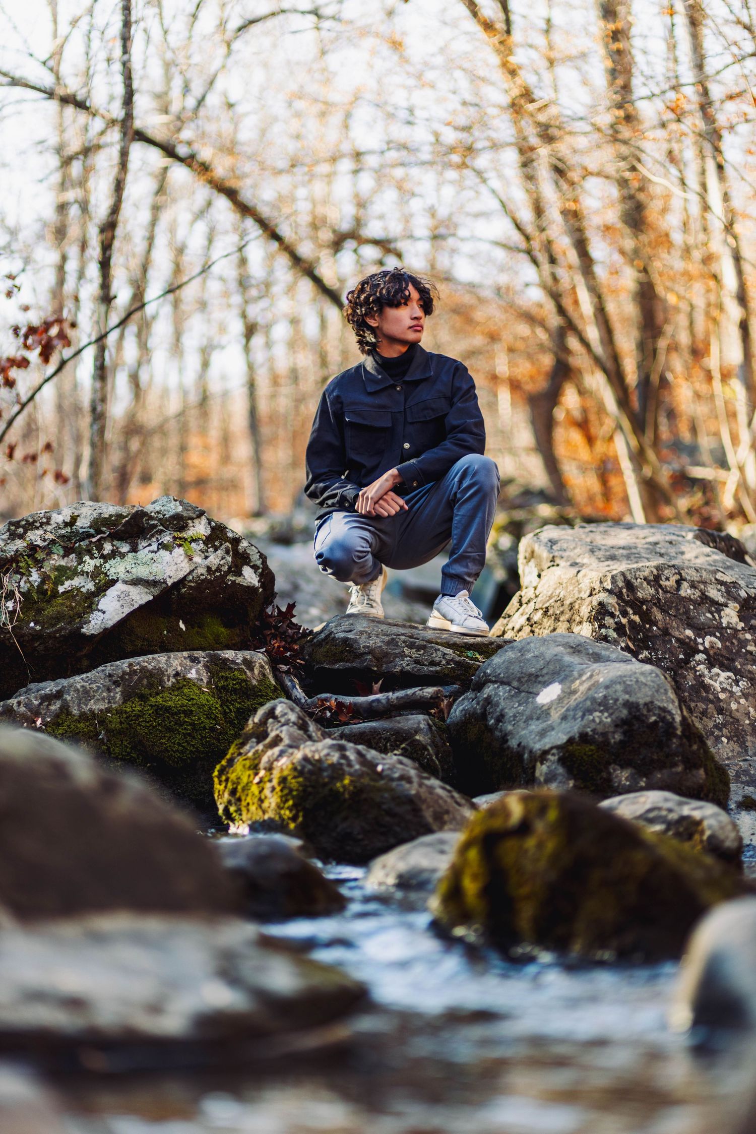 Young man crouching on mossy rocks beside forest stream in autumn woodland setting.