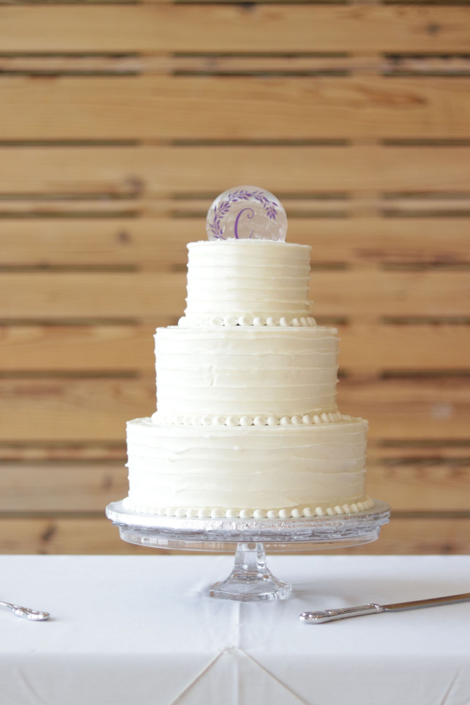 Three-tier white wedding cake with buttercream frosting and monogram topper on glass stand.