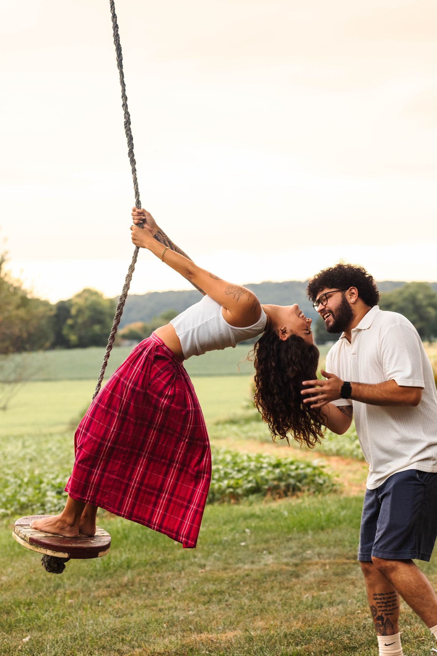 Playful couple sharing kiss as woman swings on rope swing in green field.