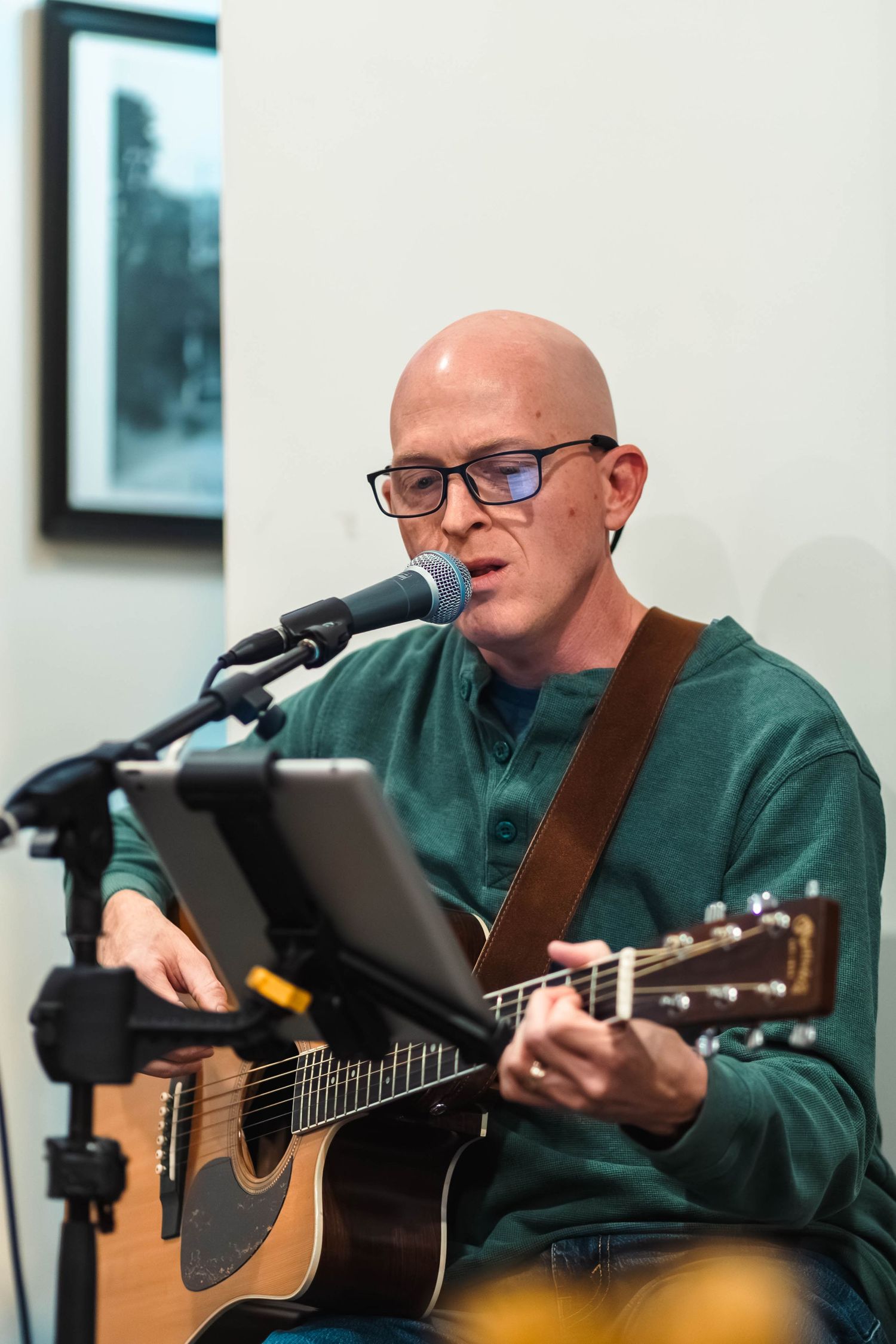 Bald man with glasses playing acoustic guitar and singing into microphone at indoor event.