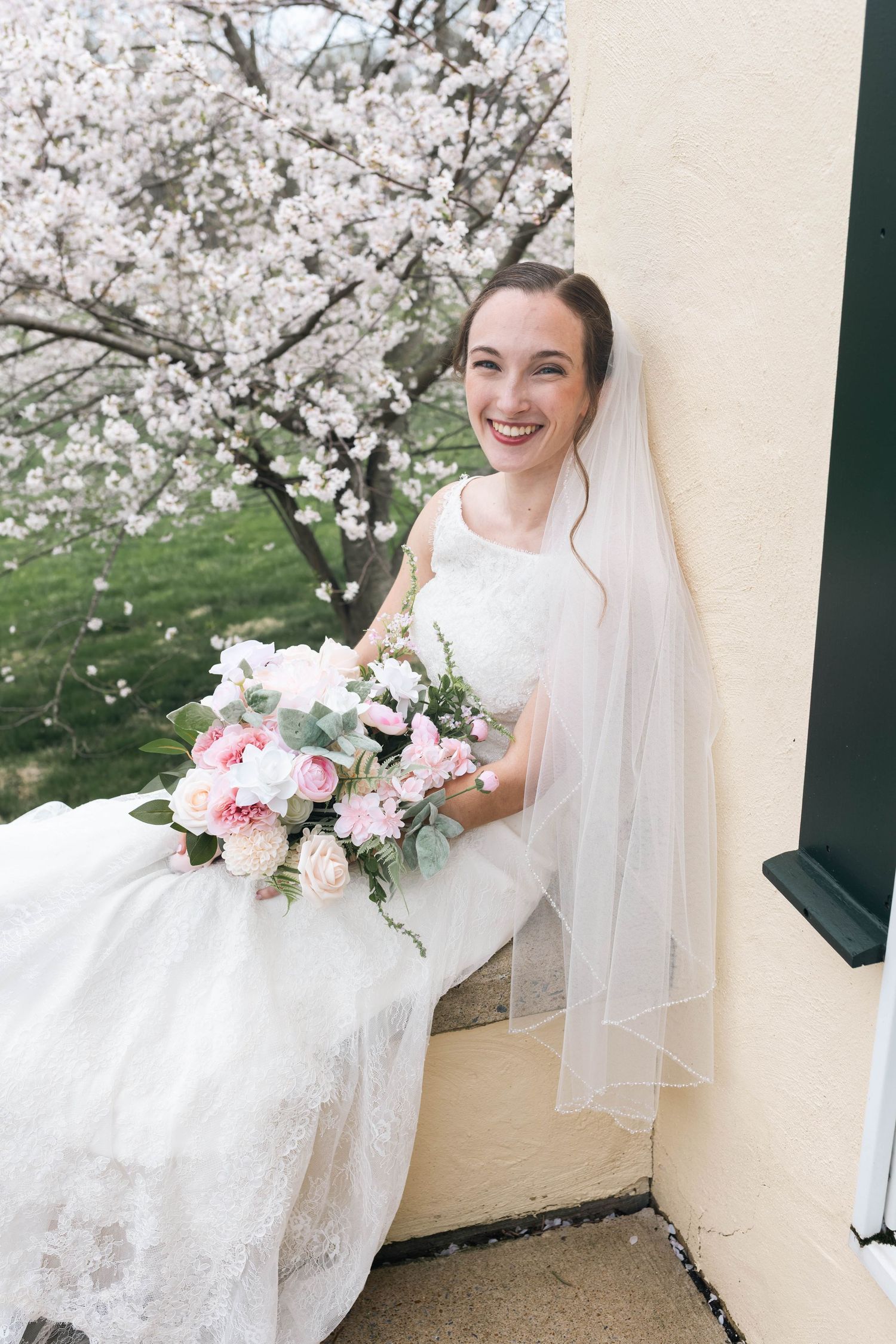 Radiant bride in lace gown and veil holding pink bouquet beside blooming cherry blossom tree.