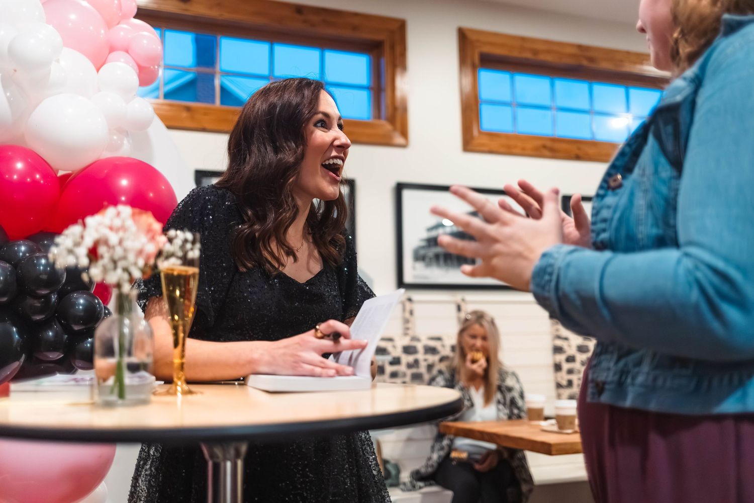 Smiling woman in black sequin dress at book signing event with pink and black balloon decorations at Brakeman's Cafe in Boyertown, PA.