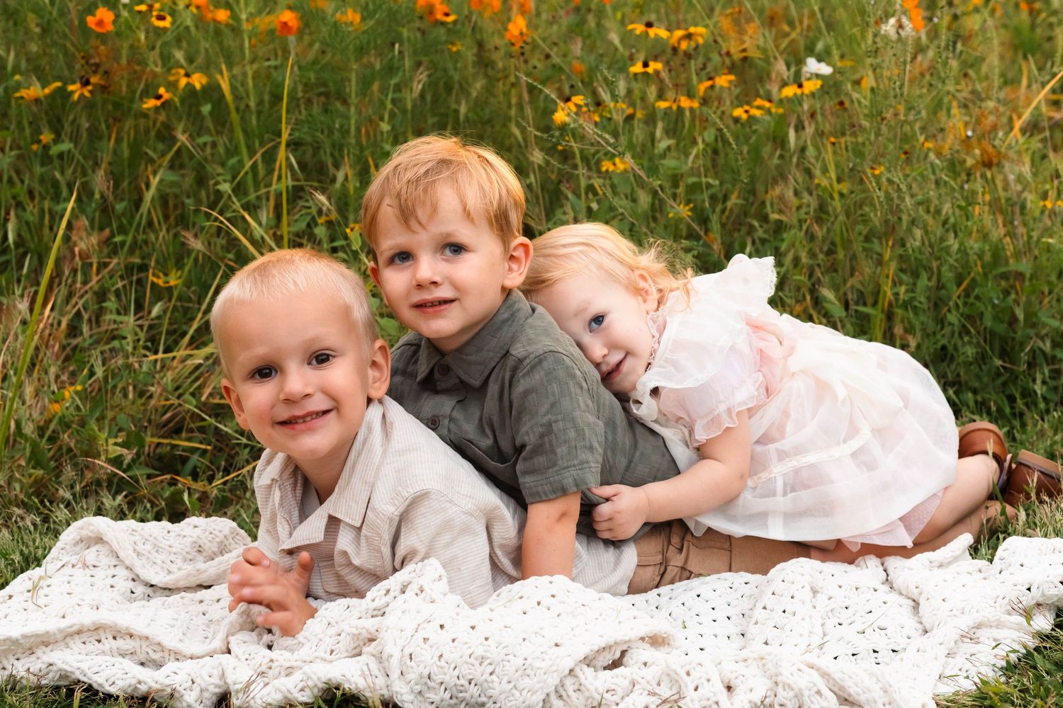 Three young siblings lying together on a white blanket in a vibrant wildflower meadow in Pekriomenville, PA.