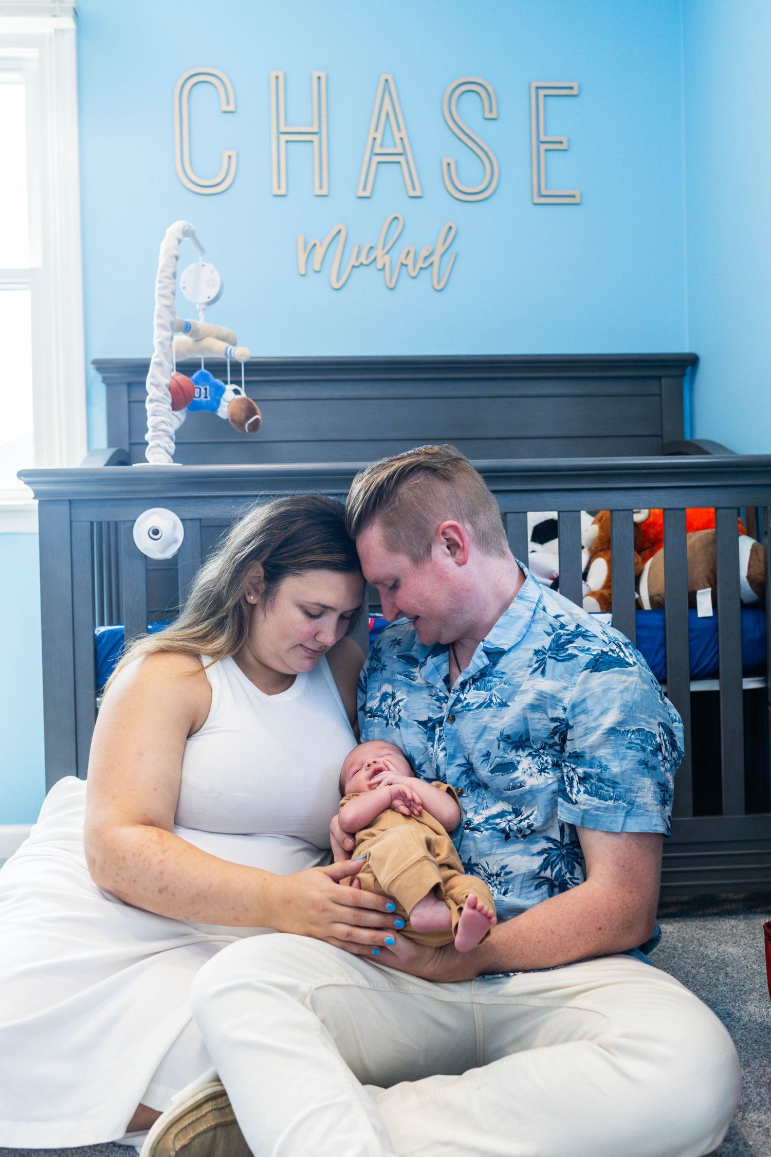 New parents sitting on nursery floor holding newborn baby Chase Michael in his blue room during an at home newborn session Philadelphia, PA.