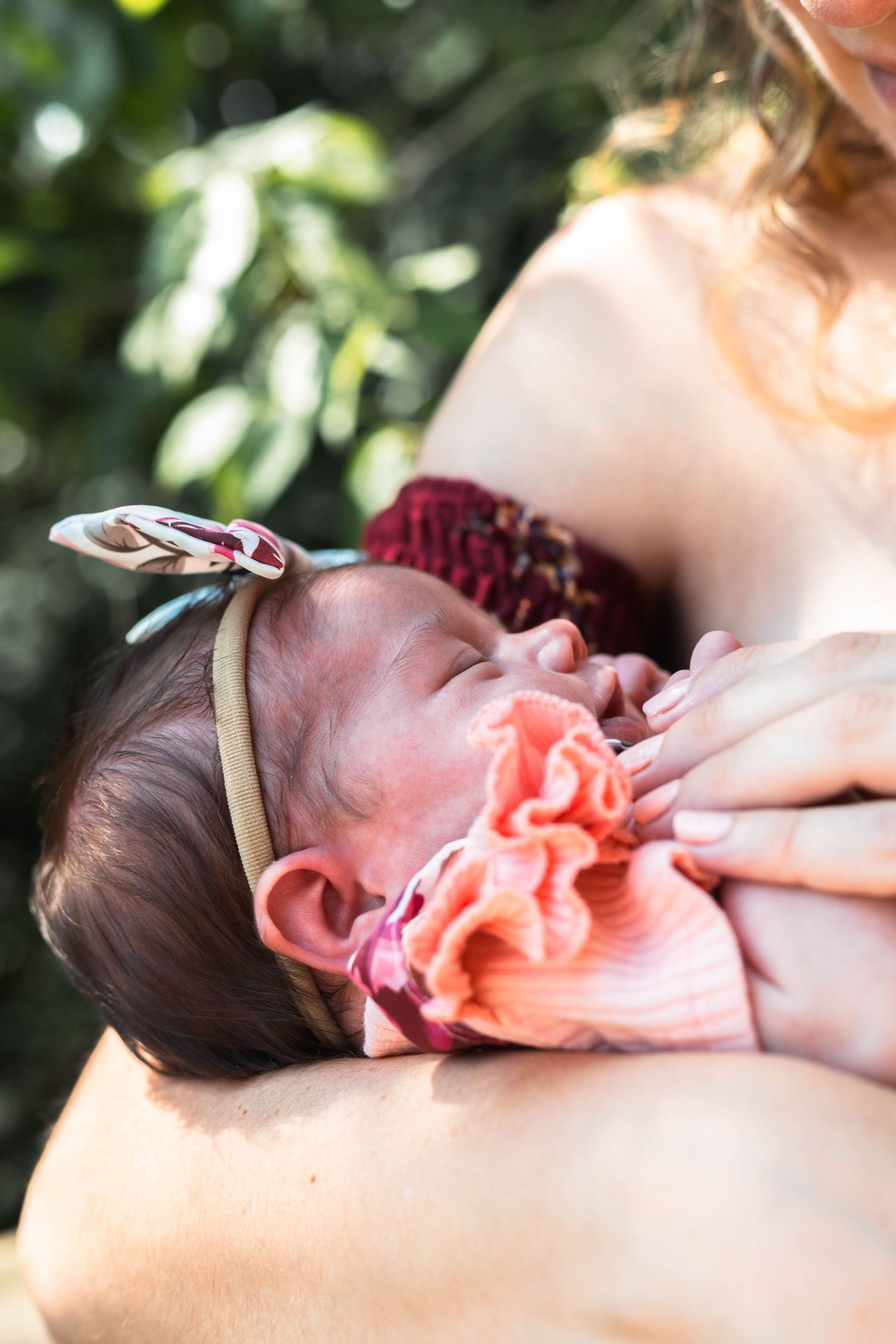 Newborn baby girl wearing a floral headband sleeping peacefully in mother's arms outdoors.