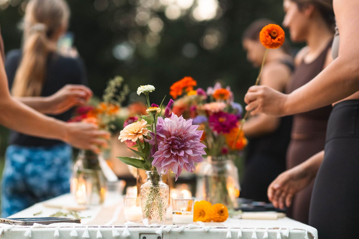 Women arrange colorful dahlia and zinnia bouquets in glass vases on a candlelit outdoor table.