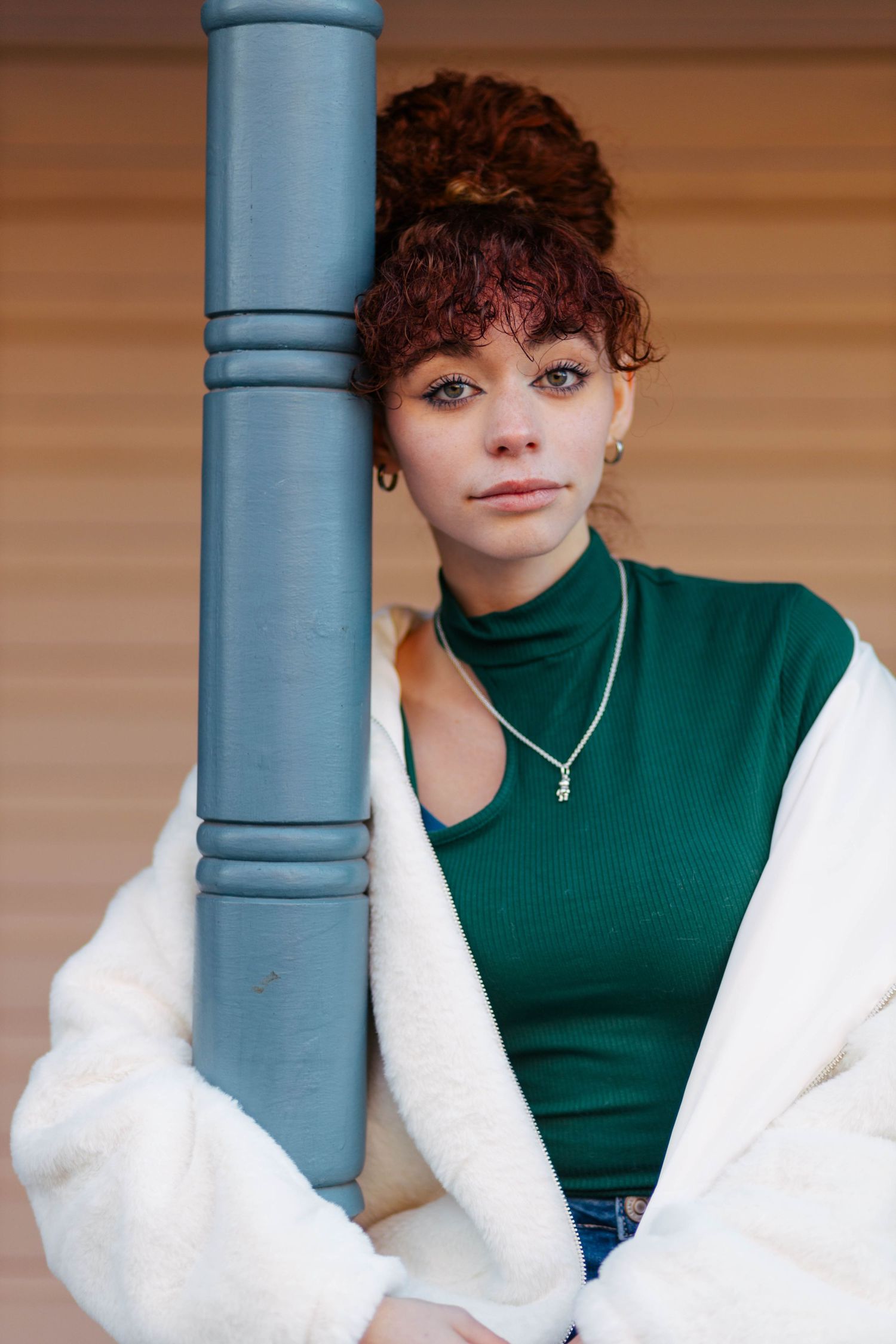 Young woman with red curly updo leaning against blue pillar in green turtleneck and white jacket.