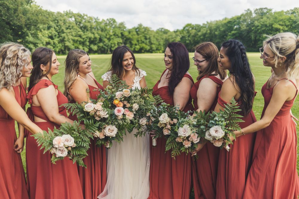 Bride surrounded by bridesmaids in rust red gowns holding lush floral bouquets outdoors.