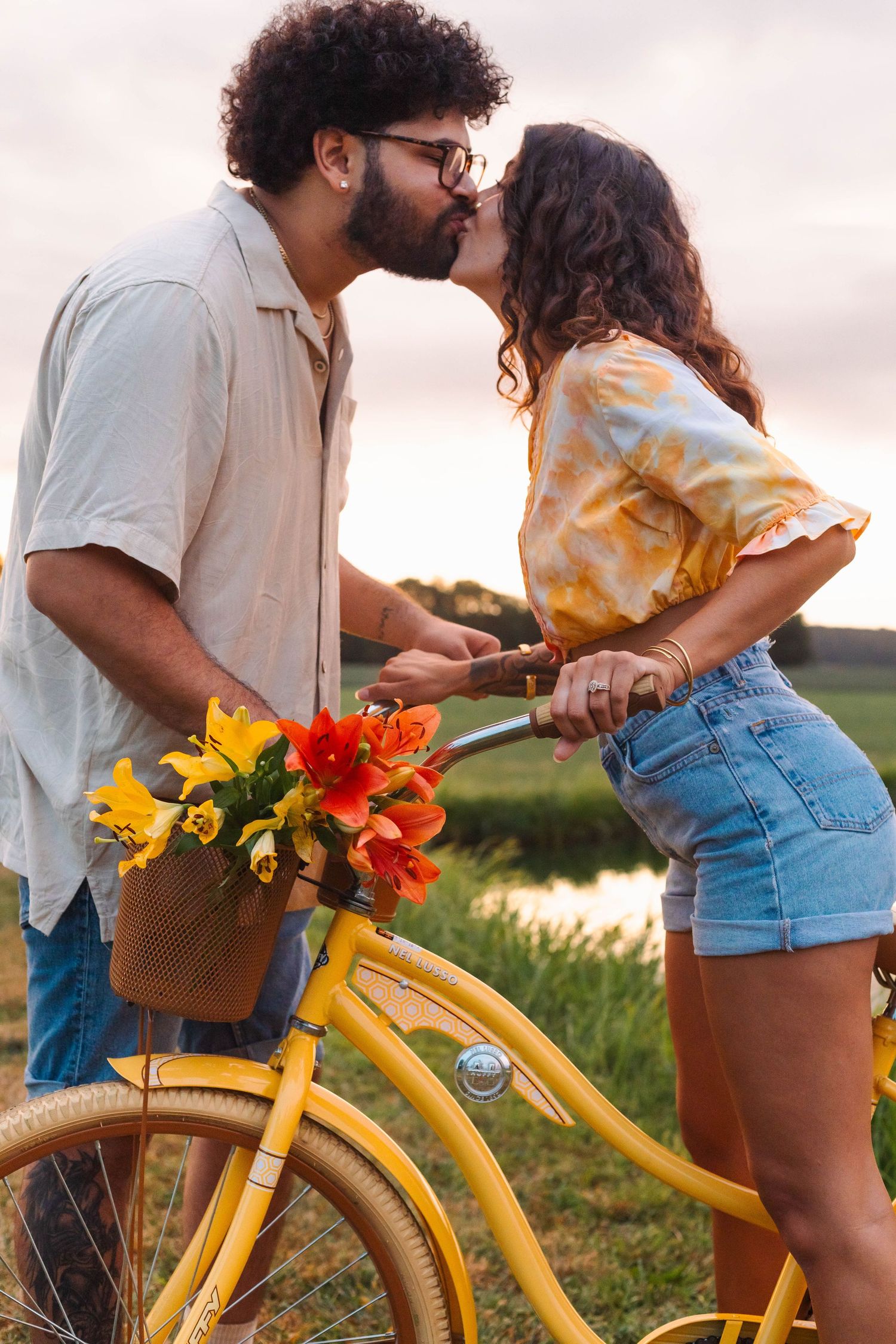 An interracial couple shares a romantic kiss beside a yellow bicycle adorned with colorful flowers at sunset in Elverson, PA.
