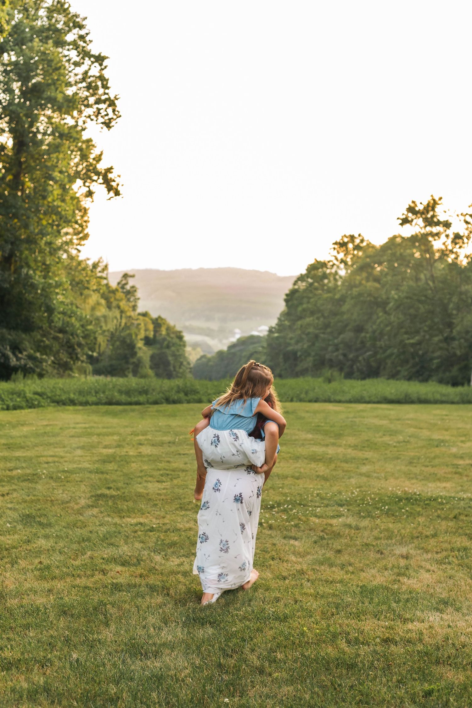 Mother carries young daughter across open green meadow at golden hour in beautiful backlit family portrait in Boyertown, PA.