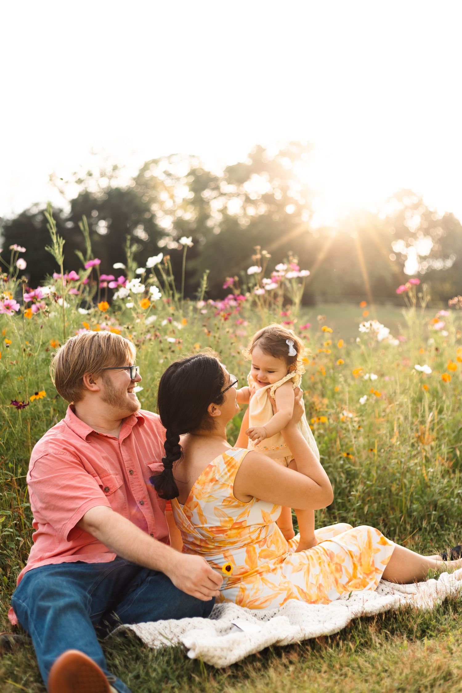 Happy family of three sitting on a blanket in a wildflower field at golden hour sunset in Pekriomenville, PA.