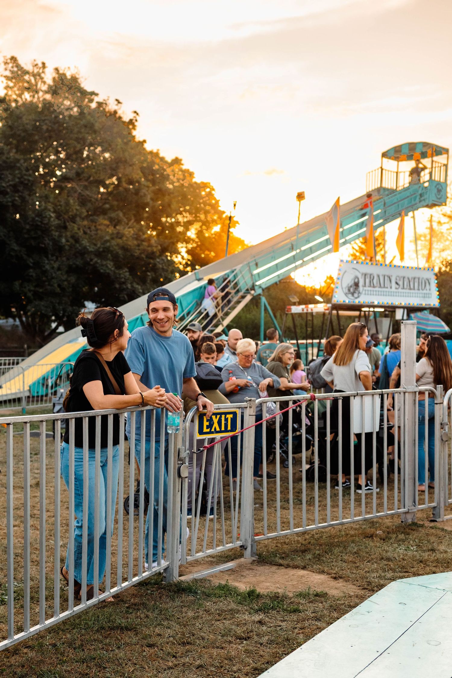 Smiling couple leaning on a metal fence at a fairground during a warm sunset evening at the Oley Fair.