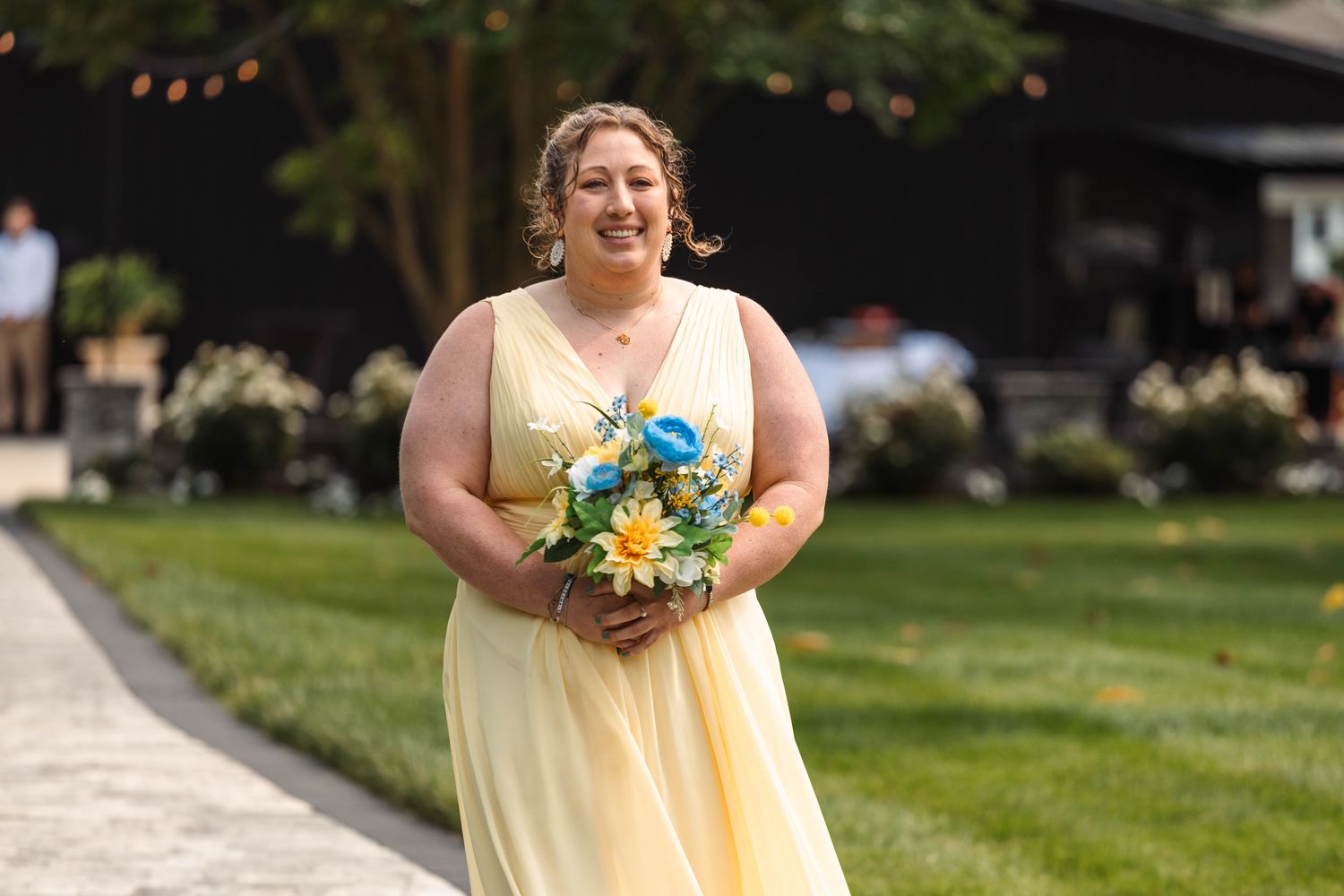 Happy bridesmaid in yellow gown holding colorful bouquet of blue and yellow flowers at outdoor wedding.