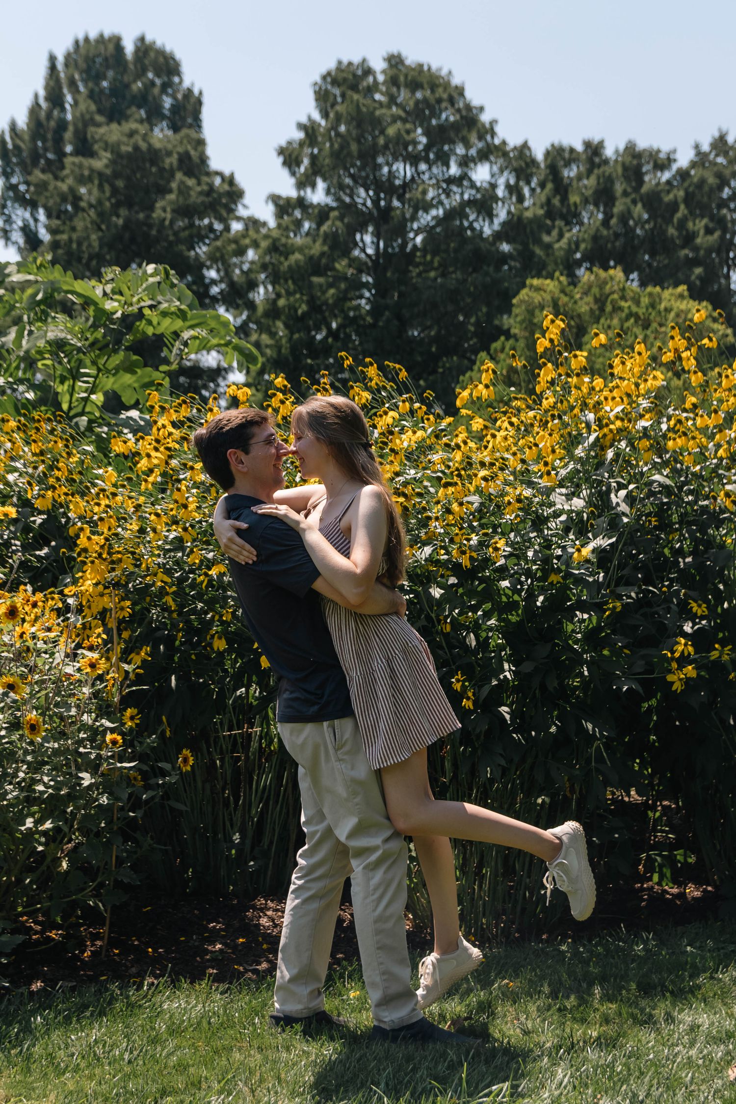 Couple embracing and smiling in front of yellow wildflowers in a lush green garden on a sunny day.