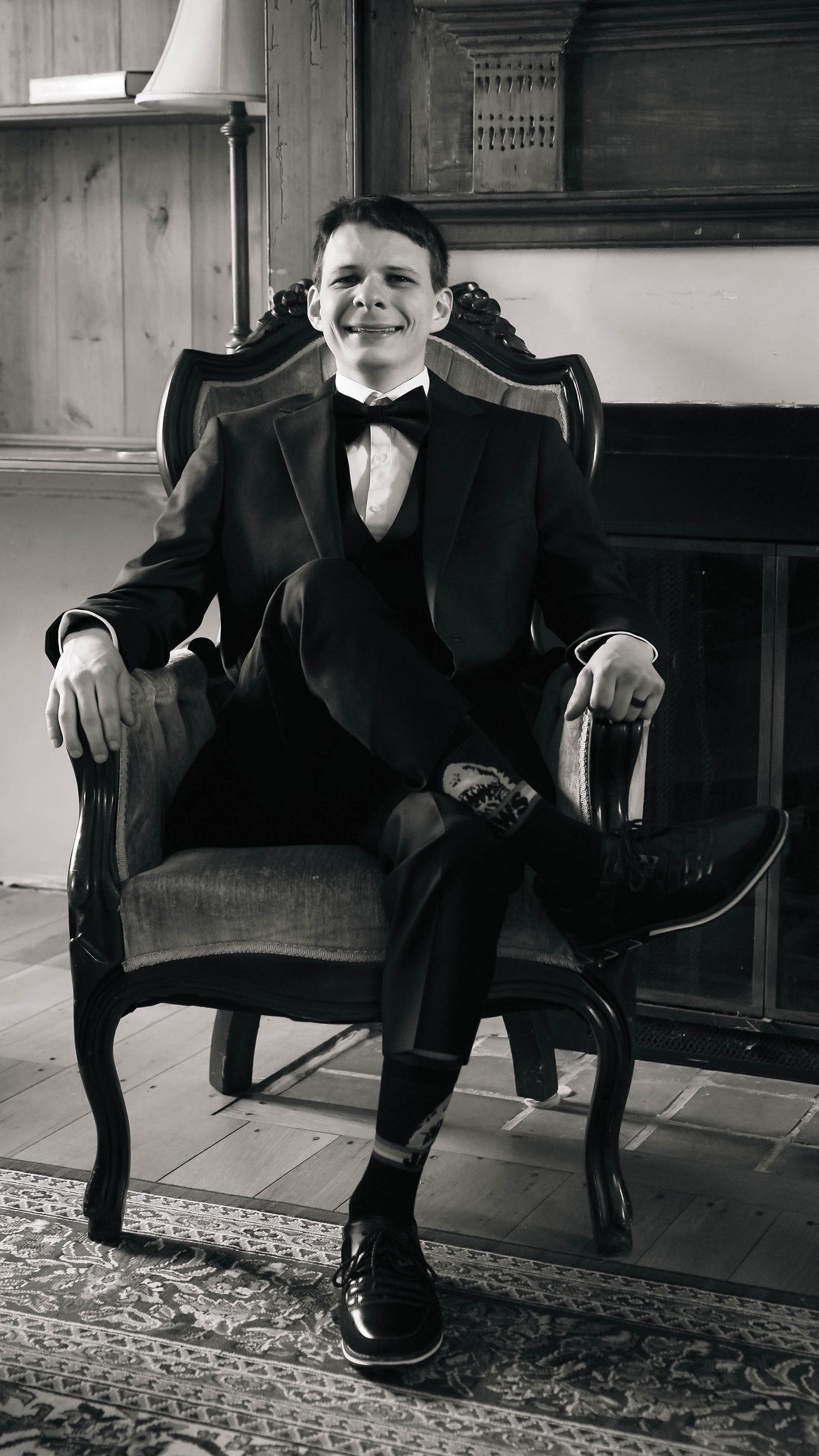Smiling groom in black tuxedo with bow tie seated in ornate chair for formal wedding portrait.