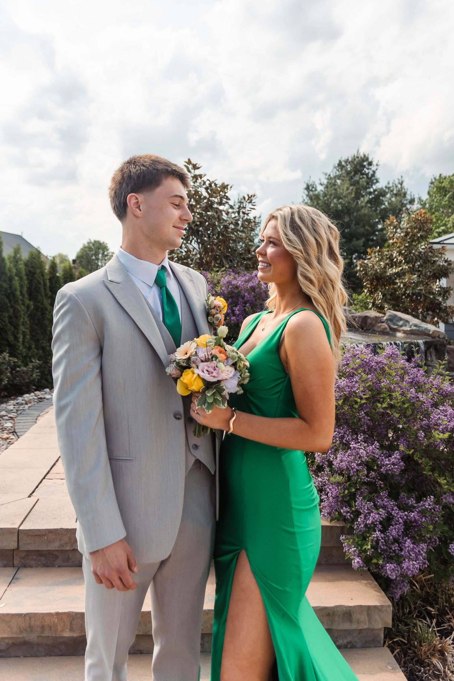 A smiling Perkiomen Valley High School couple in prom attire, a grey suit and emerald green dress, pose outdoors near purple flowers.