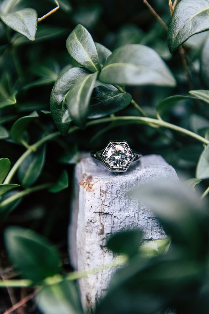 Vintage hexagonal diamond engagement ring displayed on a white stone surrounded by green leaves.