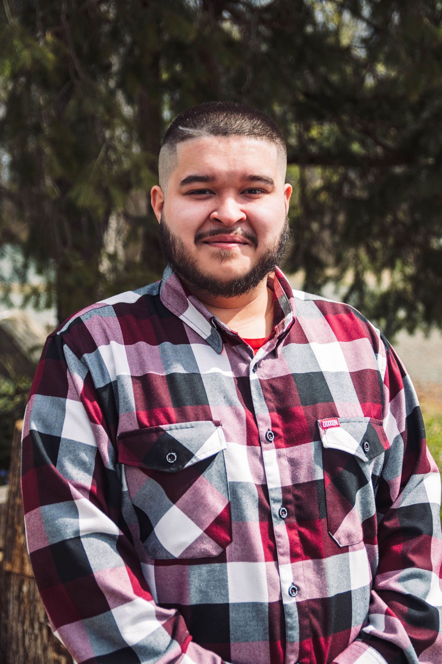 Smiling young Hispanic man with beard wearing a red plaid flannel shirt outdoors in sunlight.