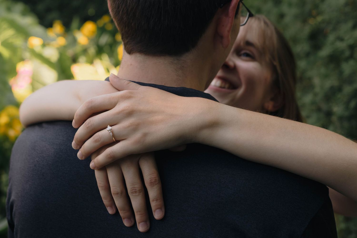 A smiling woman hugs a man from behind, showcasing an engagement ring, surrounded by lush greenery.