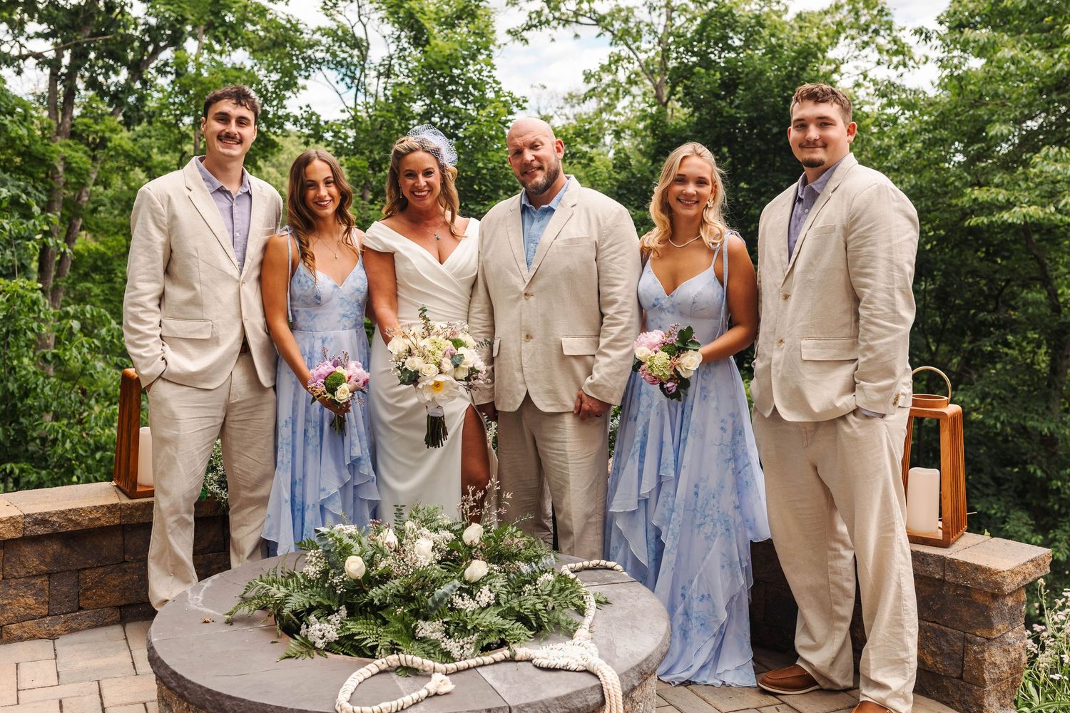 Wedding party of six posing outdoors in coordinating beige suits and blue bridesmaid dresses.