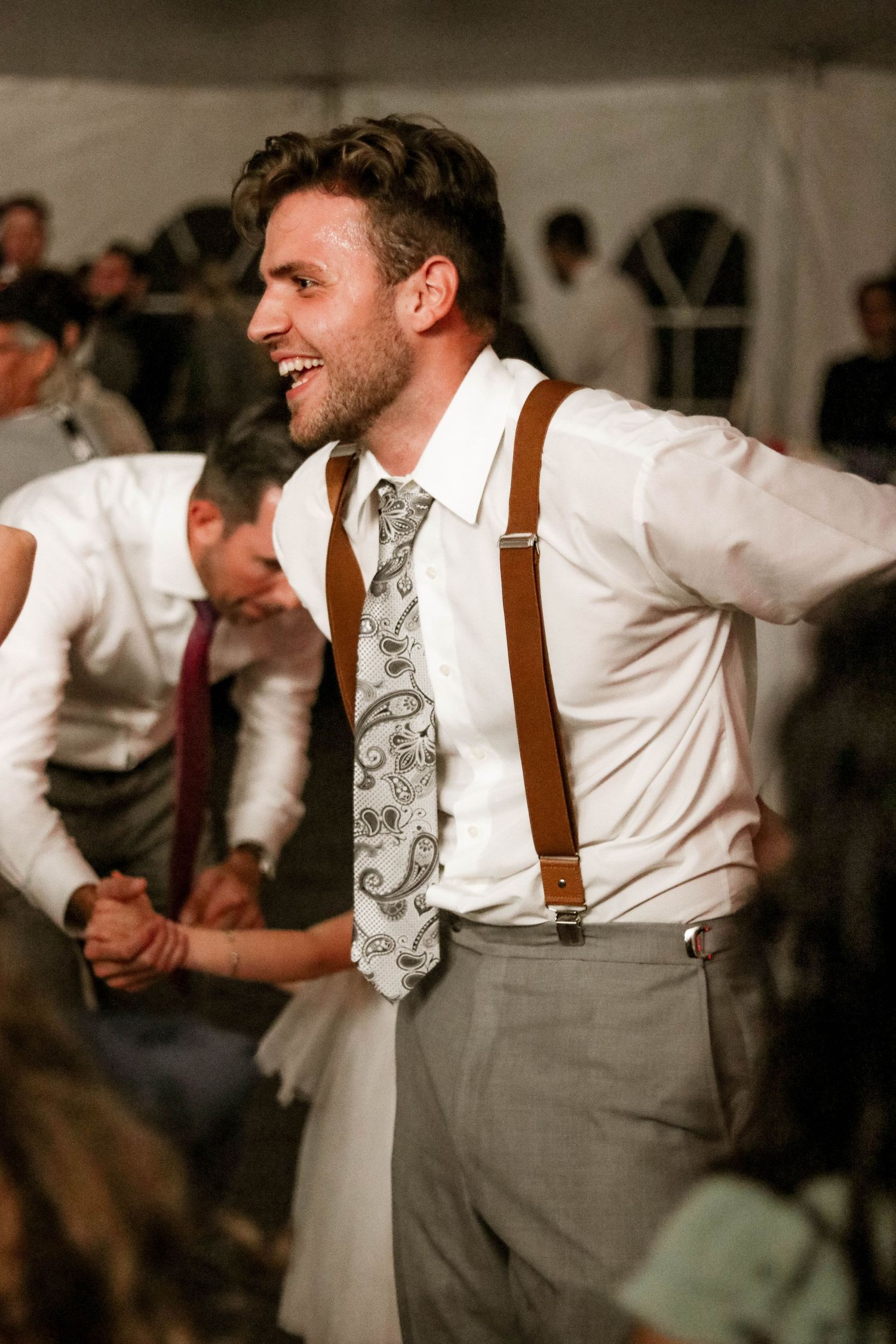 Smiling groomsman dances energetically wearing brown suspenders and paisley tie at wedding reception.