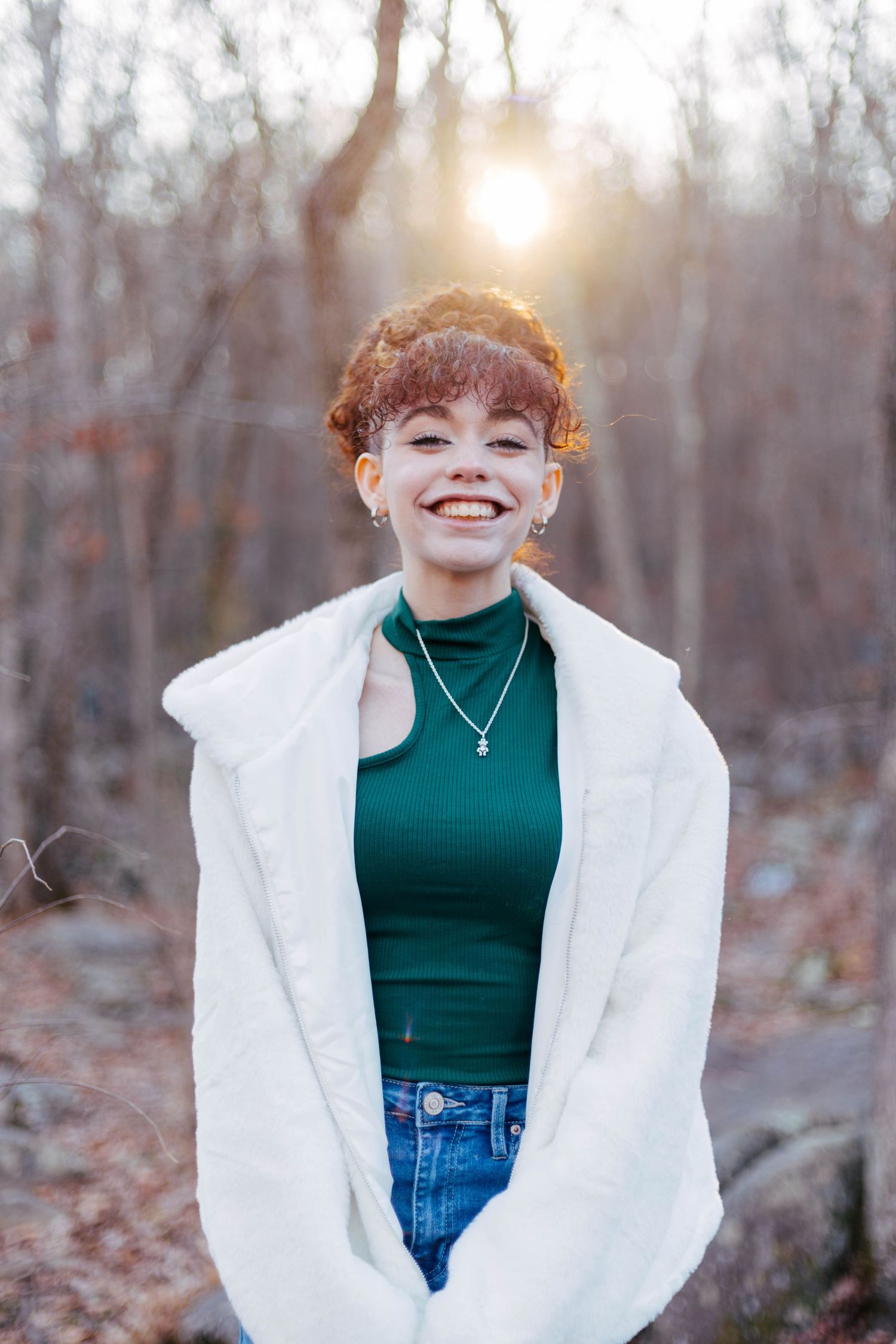 Smiling Pottstown graduate with curly red hair in green top and white fur coat posing at golden sunset.