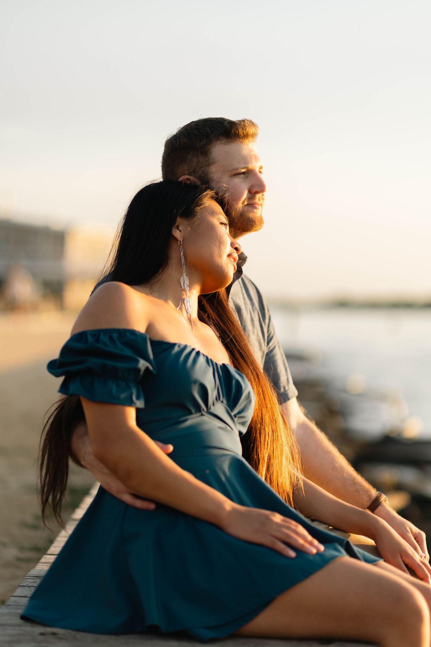 Engaged couple sits together on a beach boardwalk enjoying a golden sunset over the water in Barnegat, NJ.