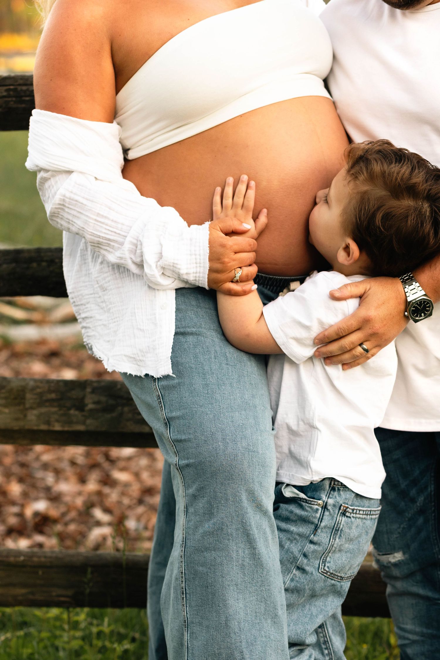 Young child lovingly kissing mother's pregnant belly while father embraces the family outdoors at Hopewell Furnace in Elverson, PA.