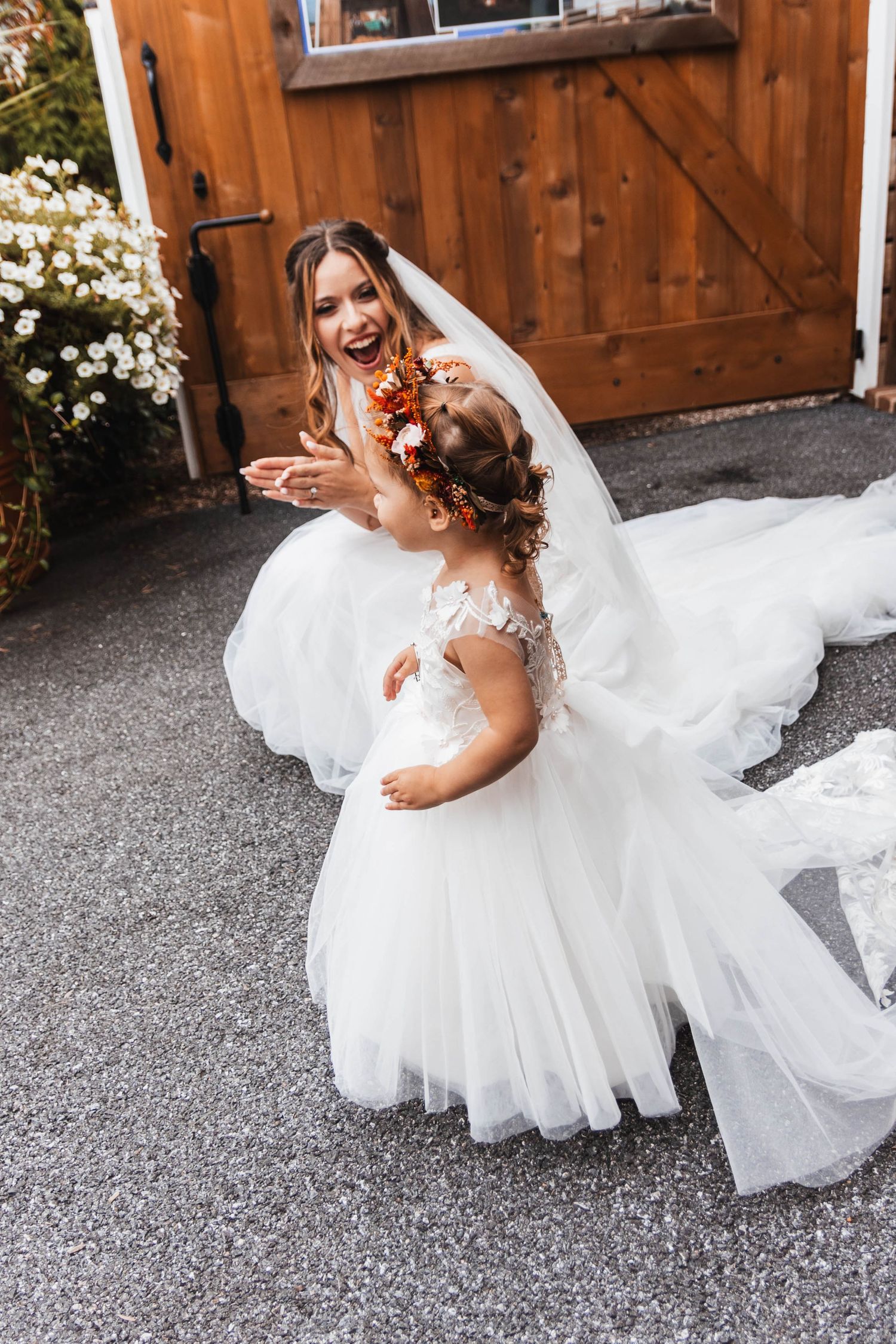 Smiling bride in lace gown shares a joyful moment with flower girl wearing an autumn floral crown.