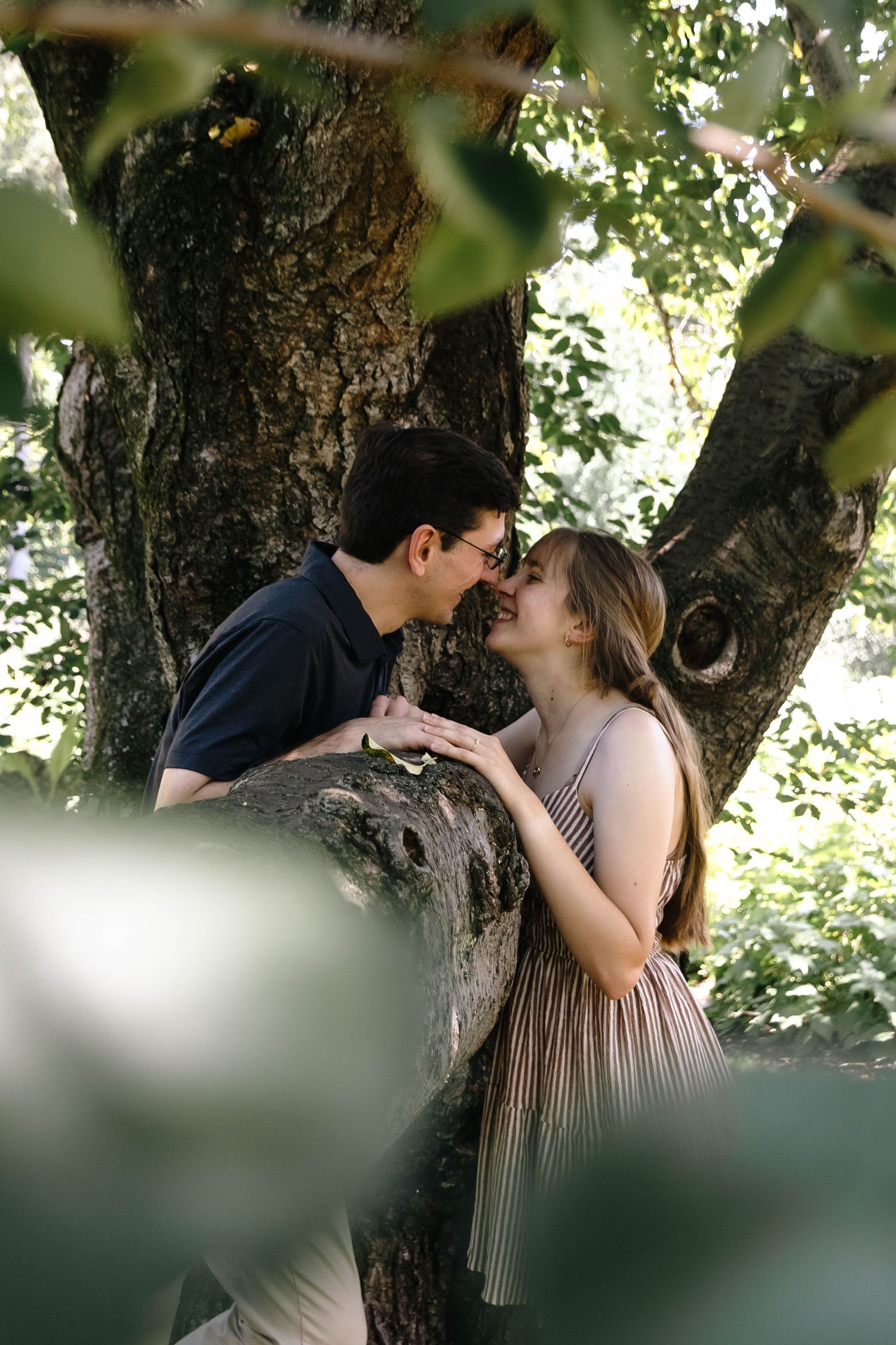 A couple shares a romantic moment leaning against a large tree trunk surrounded by lush green foliage outdoors.