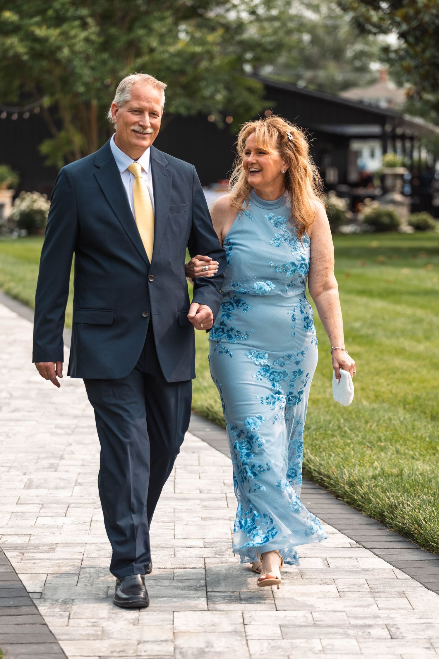 Smiling couple walking arm in arm on stone pathway dressed formally for outdoor wedding ceremony.