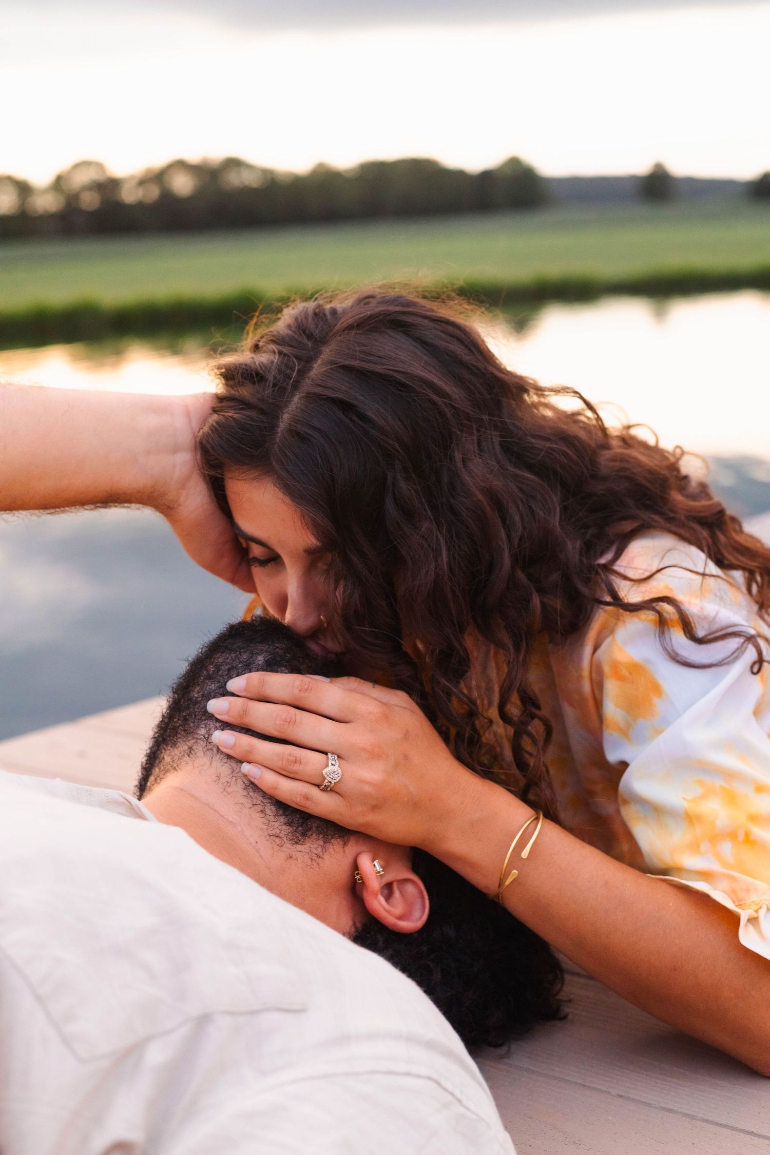 A woman with curly hair kisses a man's head while showing off her engagement ring outdoors near a lake in Elverson, PA.