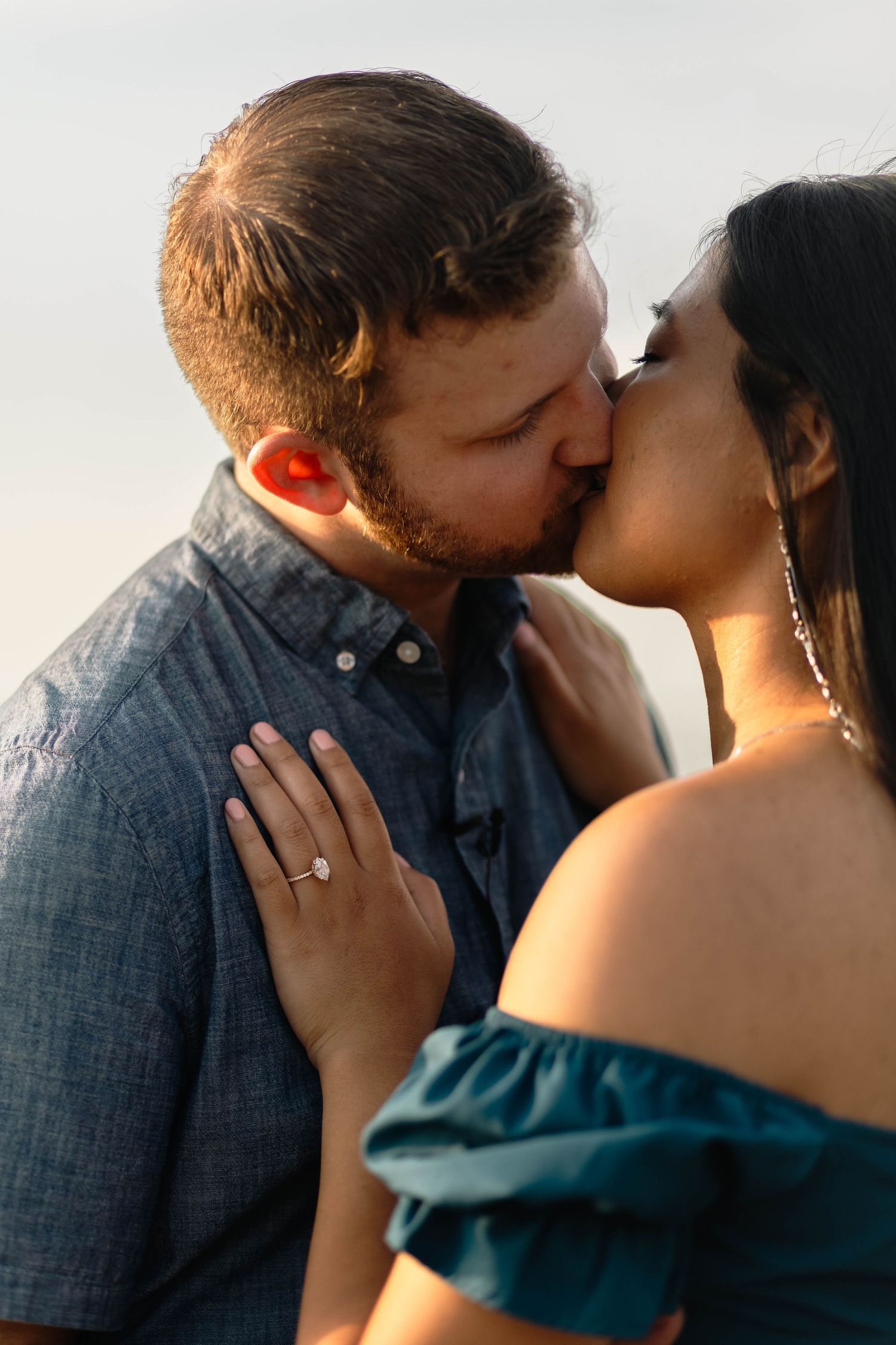 Couple shares a romantic kiss at sunset, woman displaying an engagement ring on her hand in Barnegat, NJ.