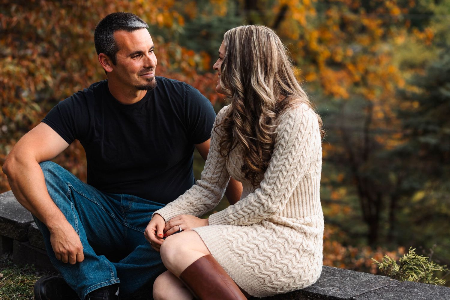 Couple sitting together on rocks in autumn park, smiling at each other in warm fall foliage setting in Boyertown, PA.