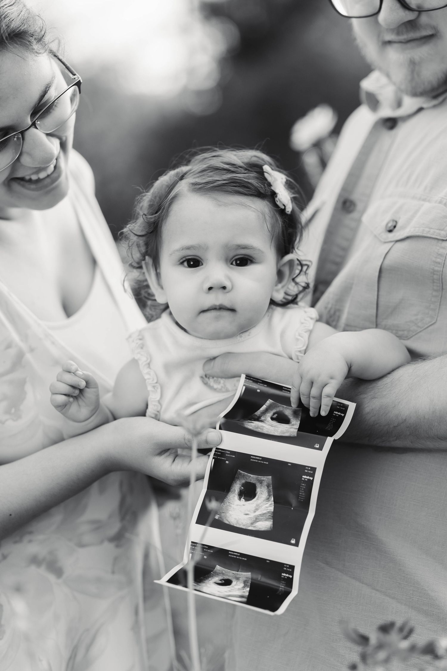 Black and white family photo of parents holding toddler girl showing ultrasound photos outdoors in Pekriomenville, PA.